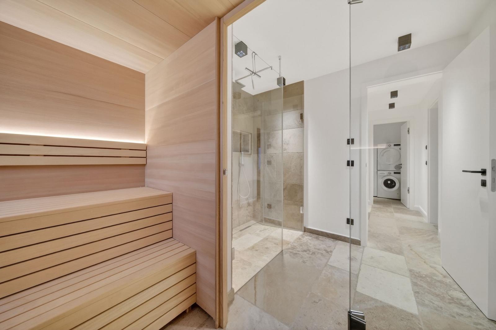 Sauna mit Holzbank, Dusche mit Glaswand und Waschraum im Hintergrund.