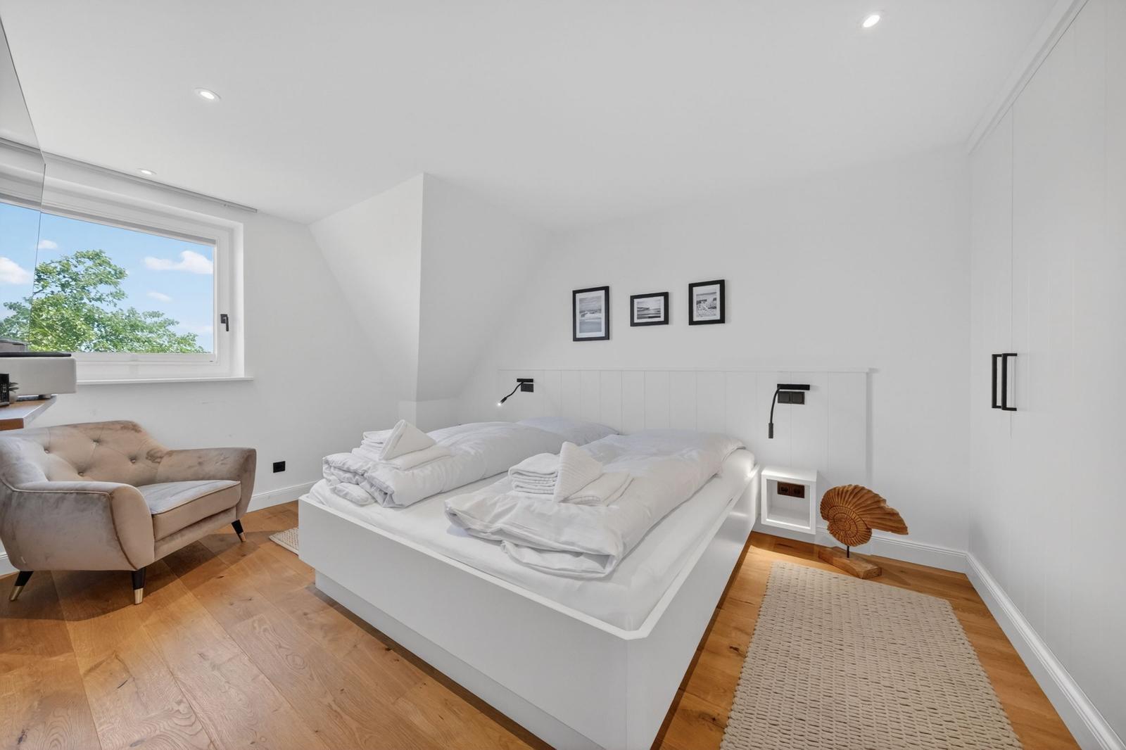 Schlafzimmer mit Doppelbett, Fenster und Sessel. Holzboden und weiße Wände.