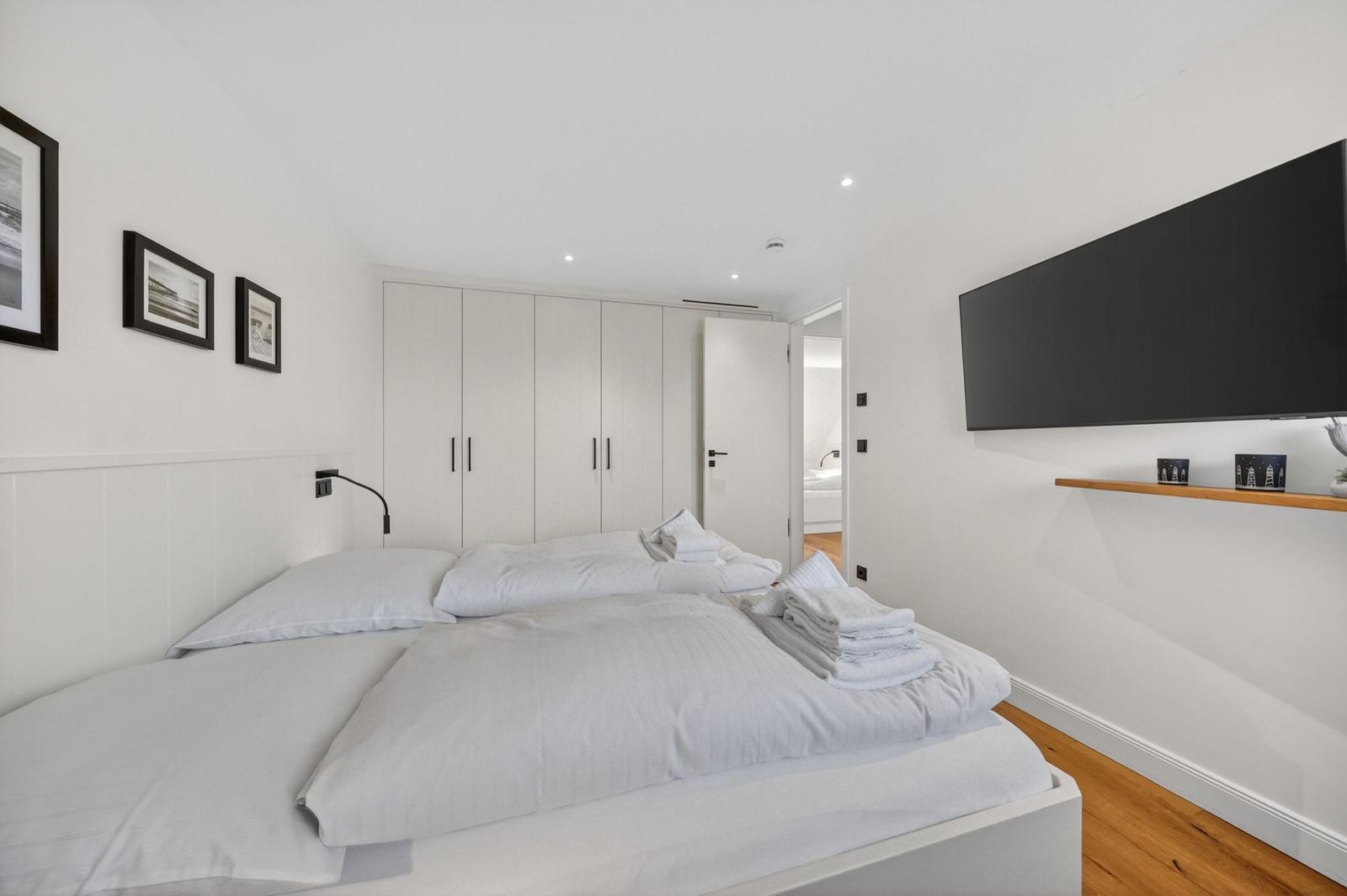 Schlafzimmer mit weißem Bett, Kleiderschrank und Wand-TV. Holzboden und Bilder an der Wand.