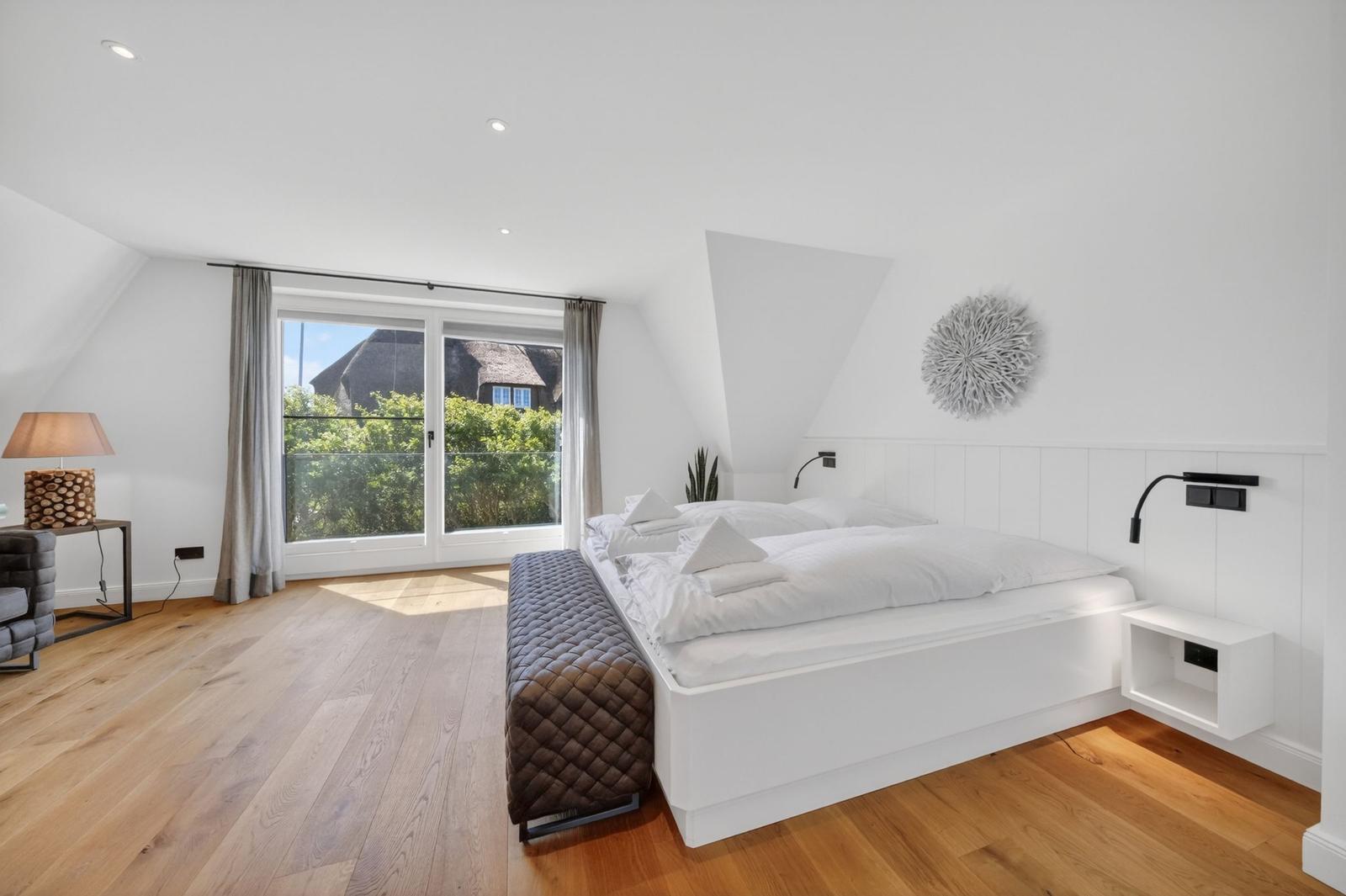 Schlafzimmer mit Dachschräge, großem Bett und Blick auf Balkon mit Grün.