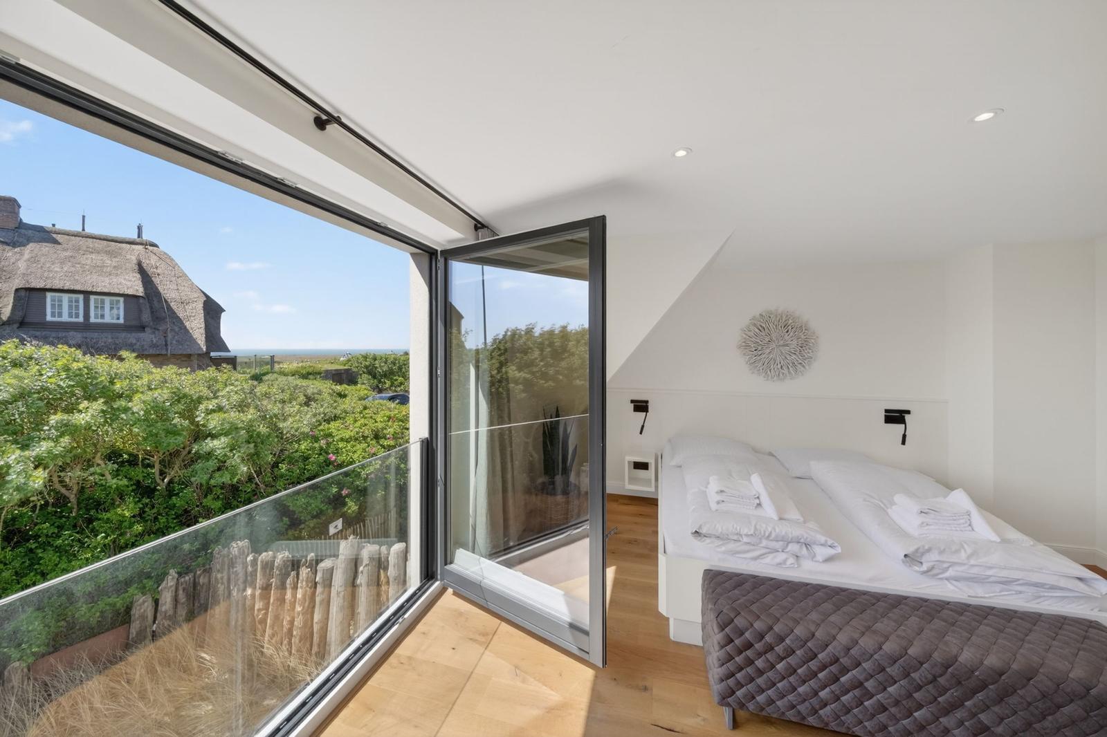 Schlafzimmer mit Blick auf Dachhütte und Grünflächen. Glasbalkon und weißes Bett.