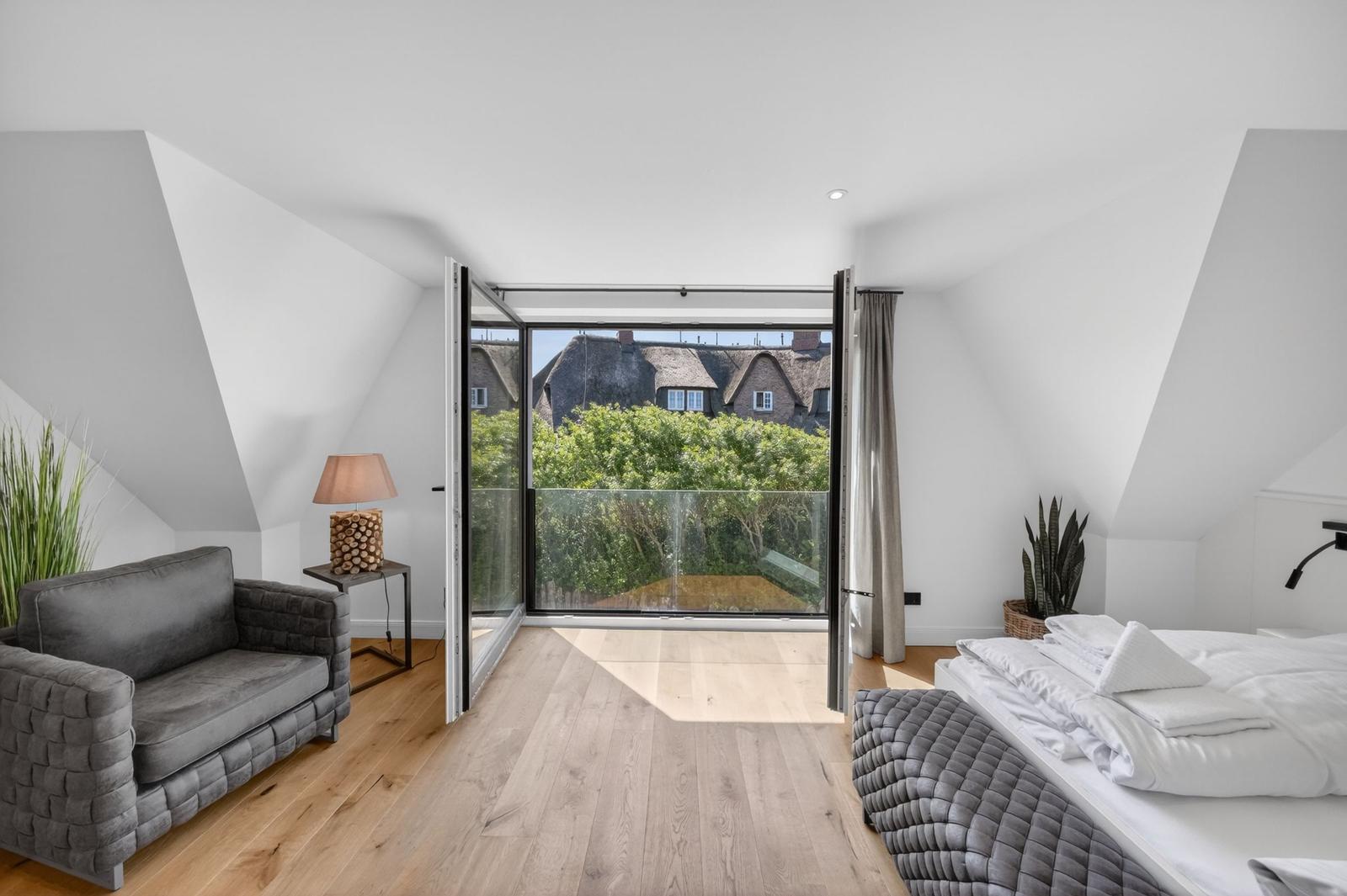 Schlafzimmer mit Balkon, Blick auf Dächer und Bäume, graue Couch, Holzboden und weißes Bett.