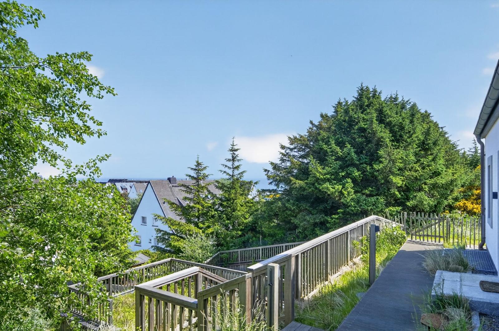 Holzterrasse mit Blick auf grüne Bäume und Ferienhaus im Hintergrund.