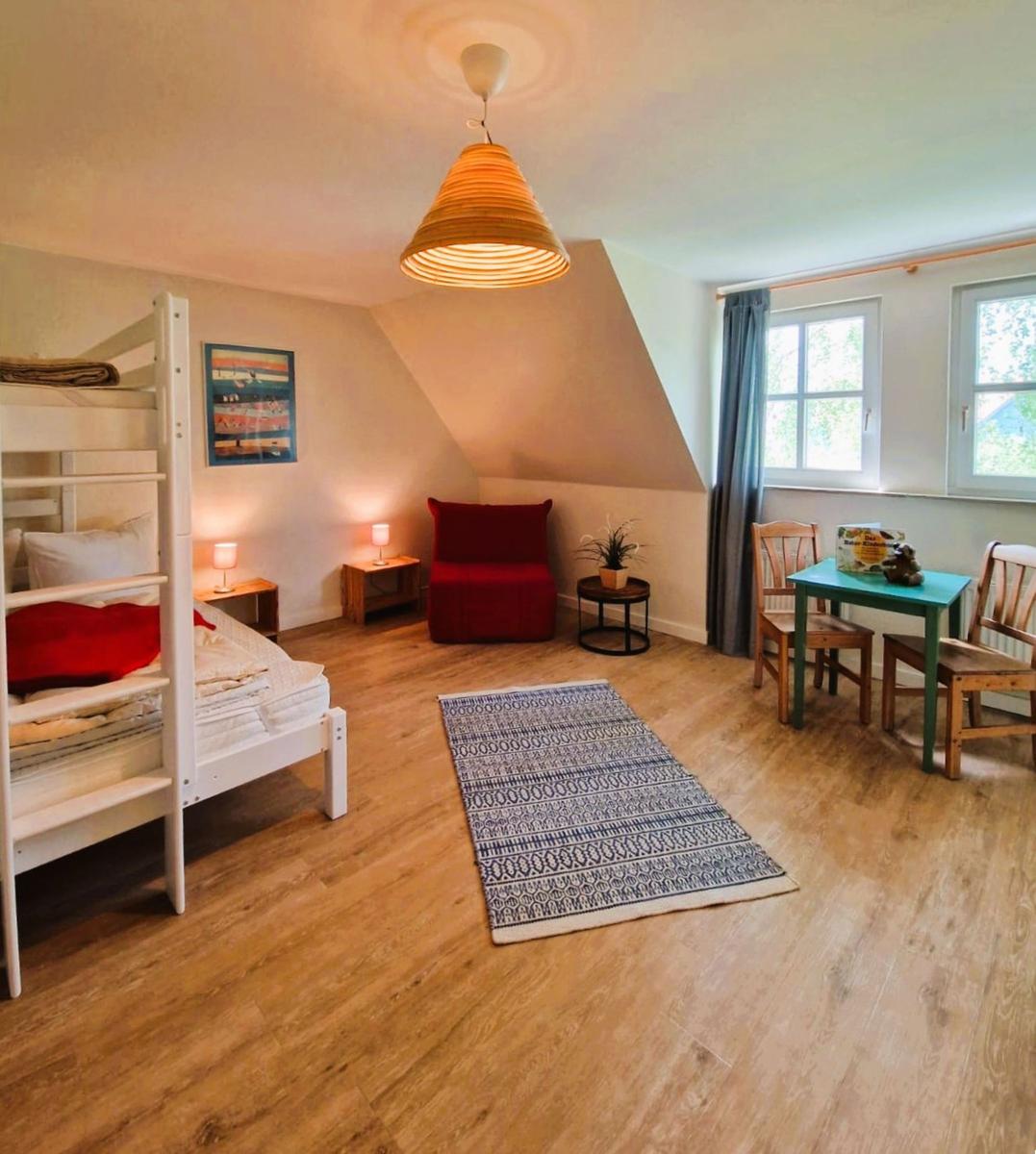 Zimmer mit Kletterbett, rotem Sessel, Tisch und Stühlen, Holzboden und Fenstern.