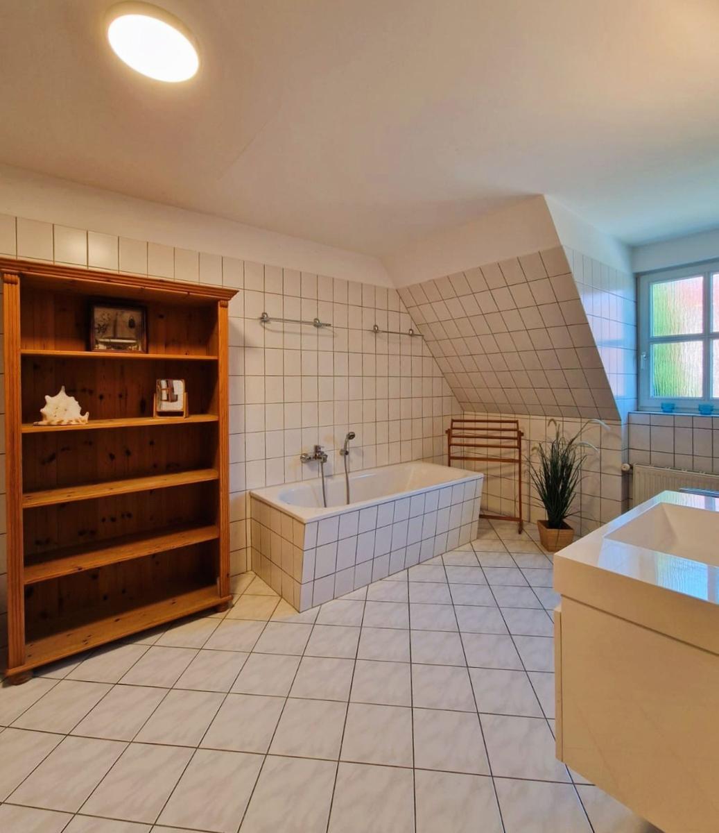 Badezimmer mit Badewanne, Holzregal und Fenster. Fliesenboden und -wände.