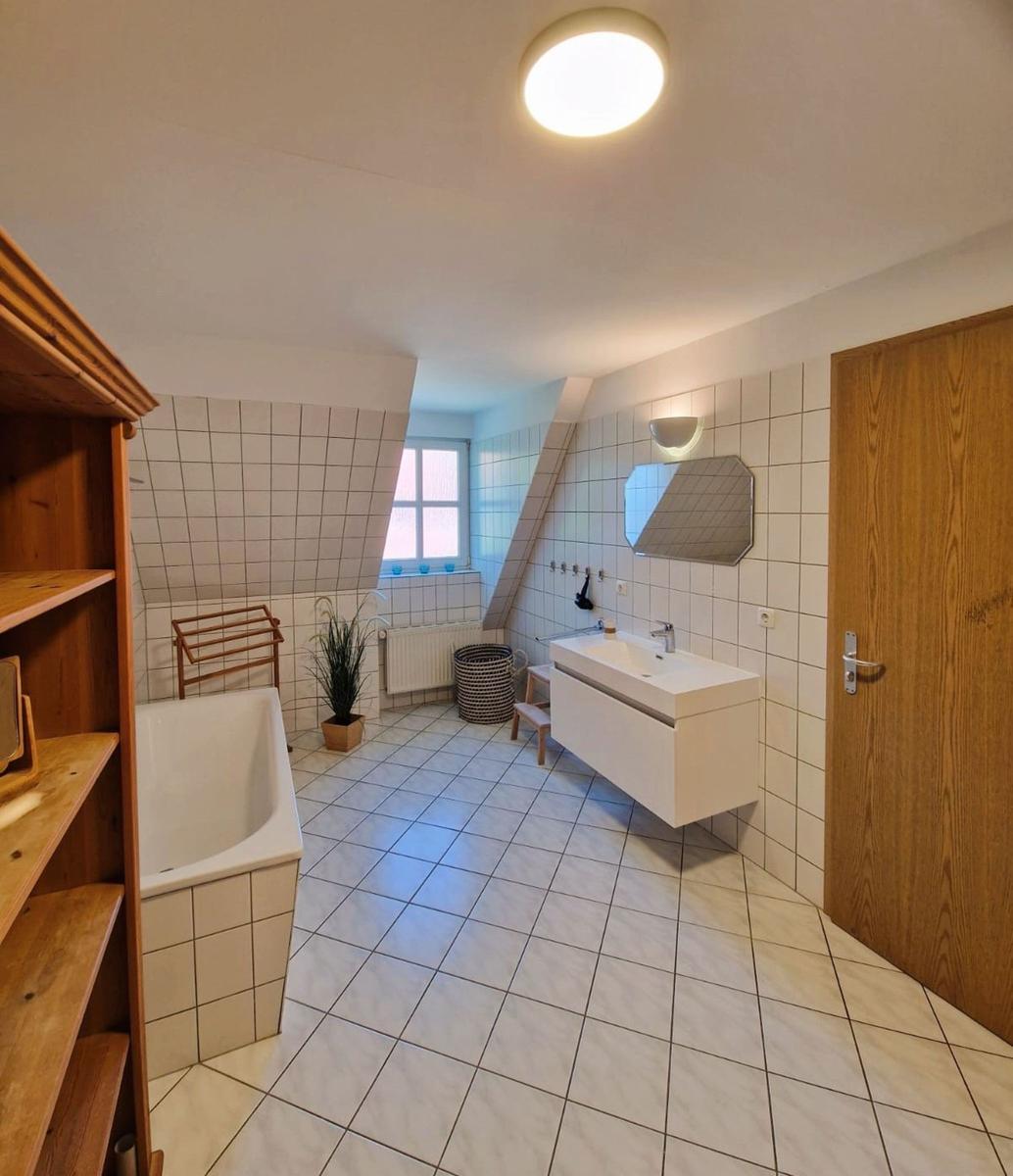 Badezimmer mit Badewanne, Waschbecken und Dusche. Holzregal, Pflanze und Fenster sichtbar.
