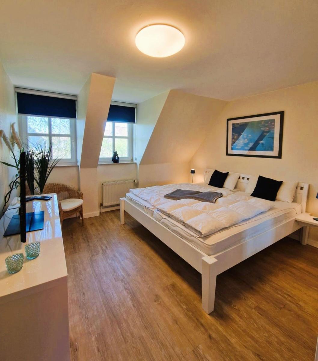 Schlafzimmer mit Doppelbett, zwei Fenstern und Holzboden. Wandbild und Schreibtisch sind vorhanden.