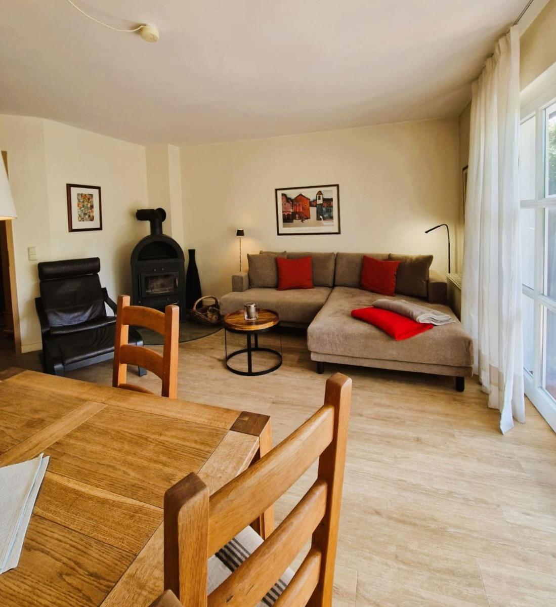 Wohnzimmer mit Sofa, Esstisch und Kaminofen. Holzfußboden und Fenster mit Vorhängen.