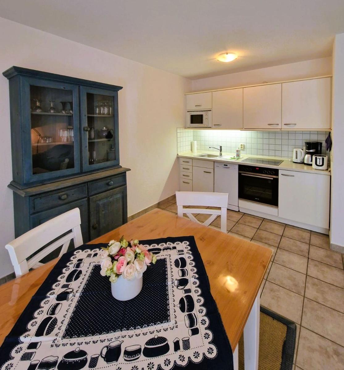 Küche mit Essbereich, weißen Schränken und blauem Sideboard. Tisch mit Tischdecke und Blumen.
