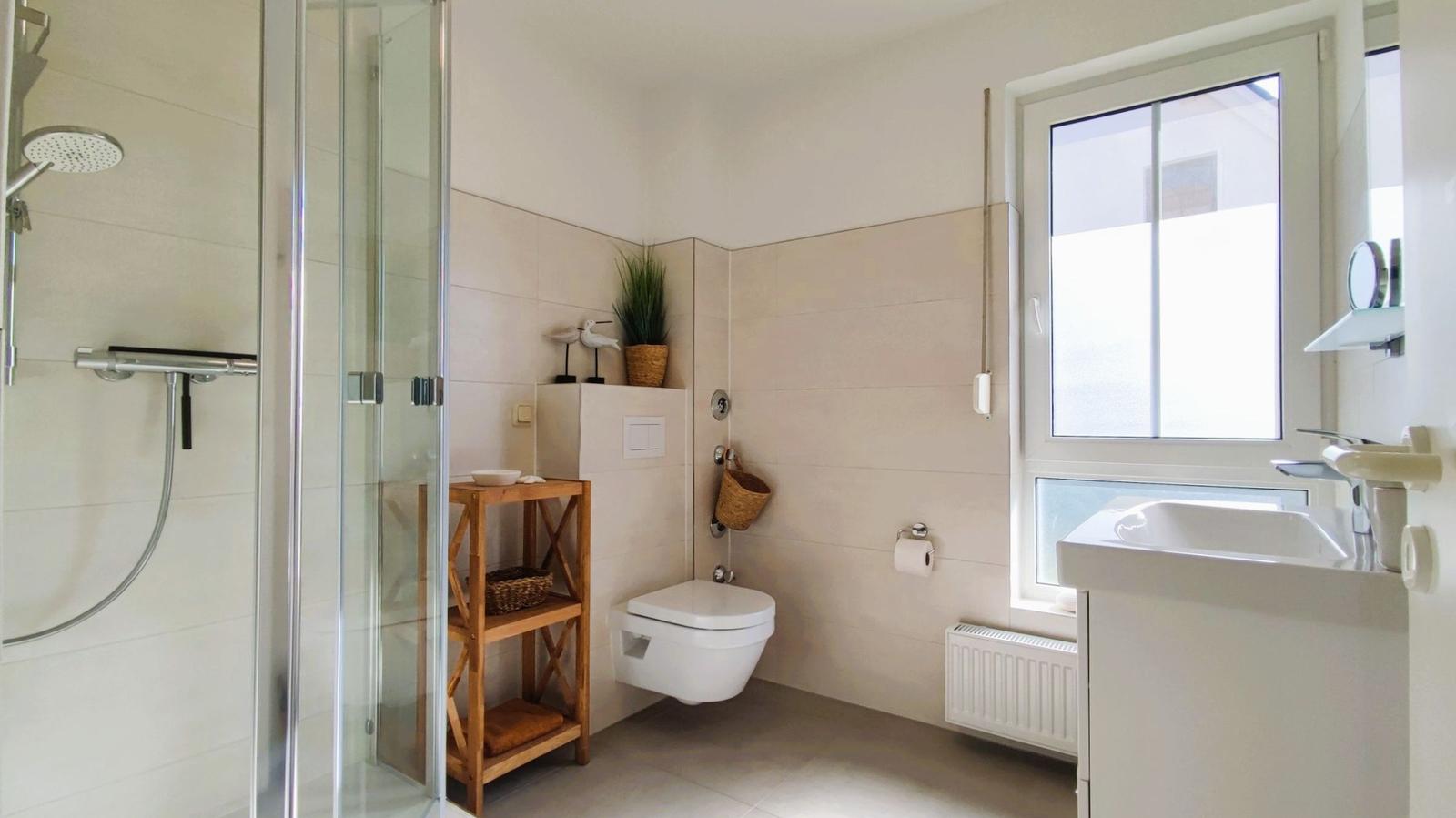 Badezimmer mit Dusche, WC und Waschbecken. Fenster und Holzregal sichtbar.