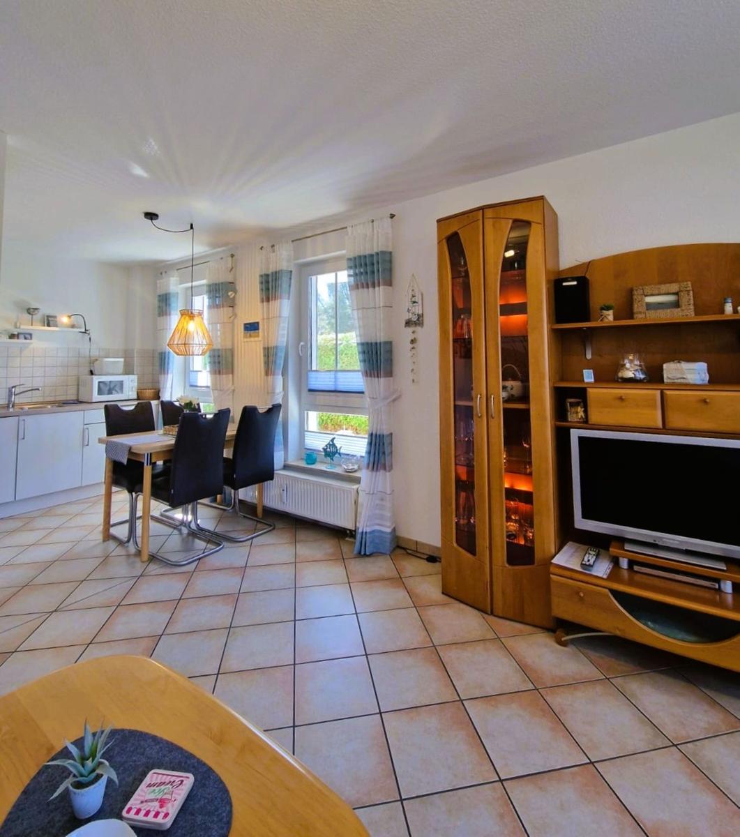 Wohn- und Essbereich mit Küchenzeile, TV und Glasvitrine. Fliesenboden und Fenster mit Vorhängen.