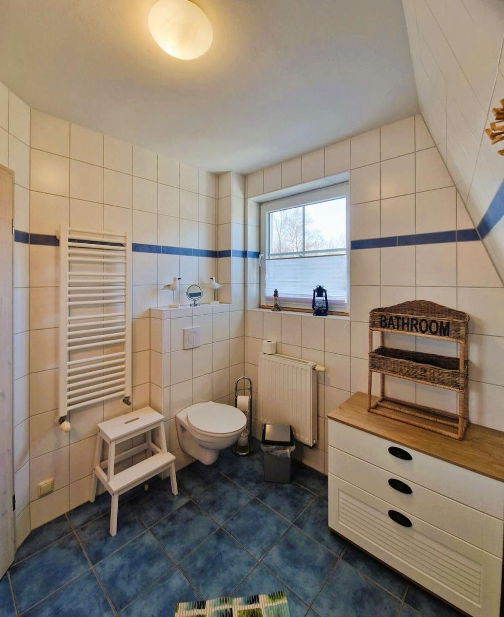Badezimmer mit Toilette, Heizkörper und Fenster. Fliesenboden und weiße Wandfliesen.