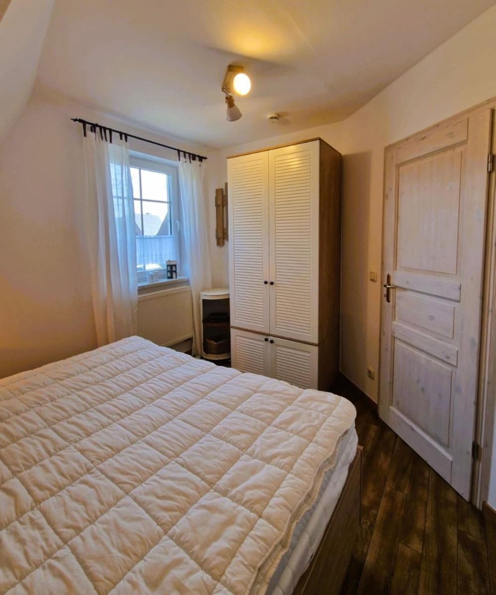 Schlafzimmer mit Bett, Kleiderschrank und Fenster mit weißen Vorhängen.