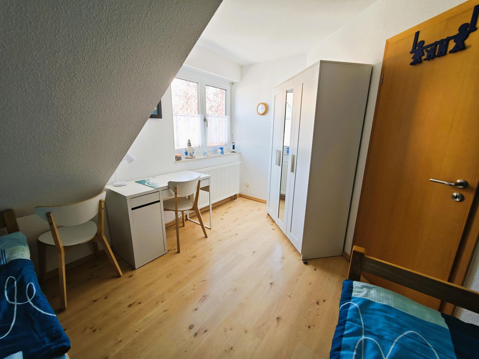 Schlafzimmer mit Schreibtisch, Kleiderschrank und Fenster. Holzboden und Tür mit Aufkleber.
