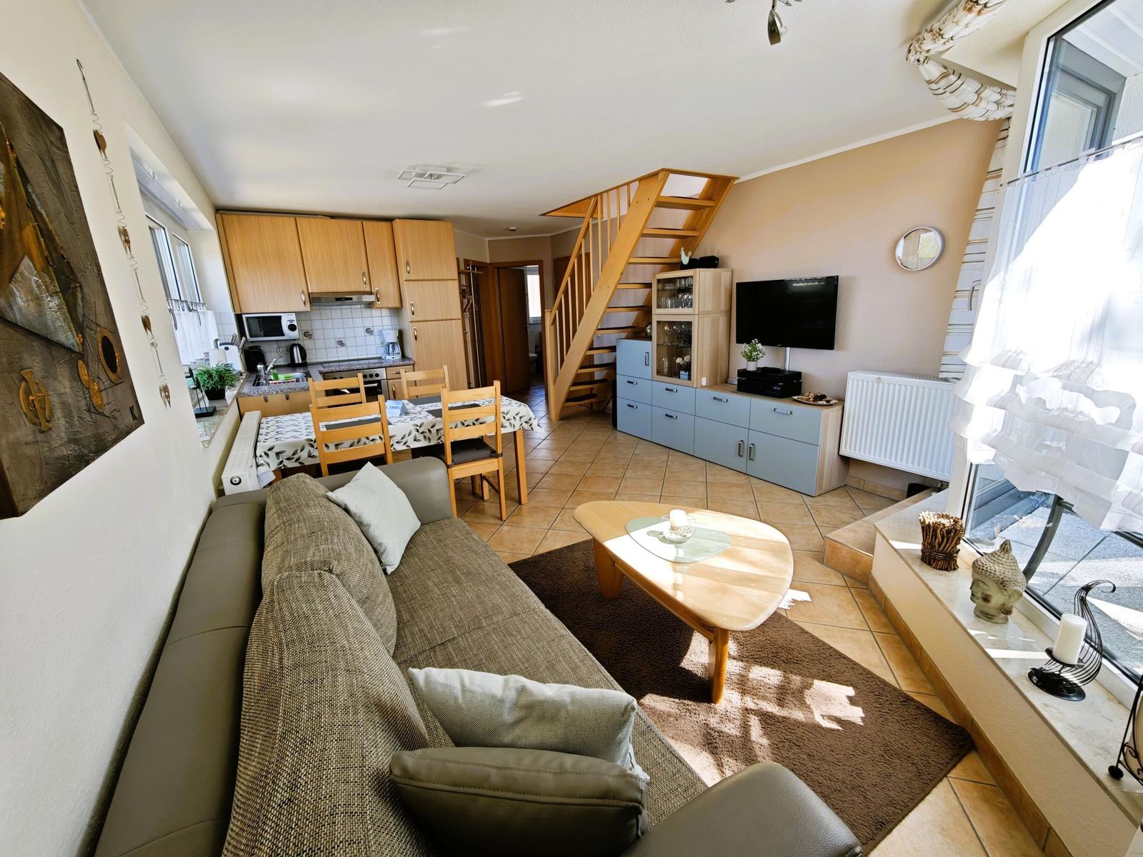 Wohnzimmer mit Sofa, Esstisch und Küchenbereich. Holztreppe führt nach oben.
