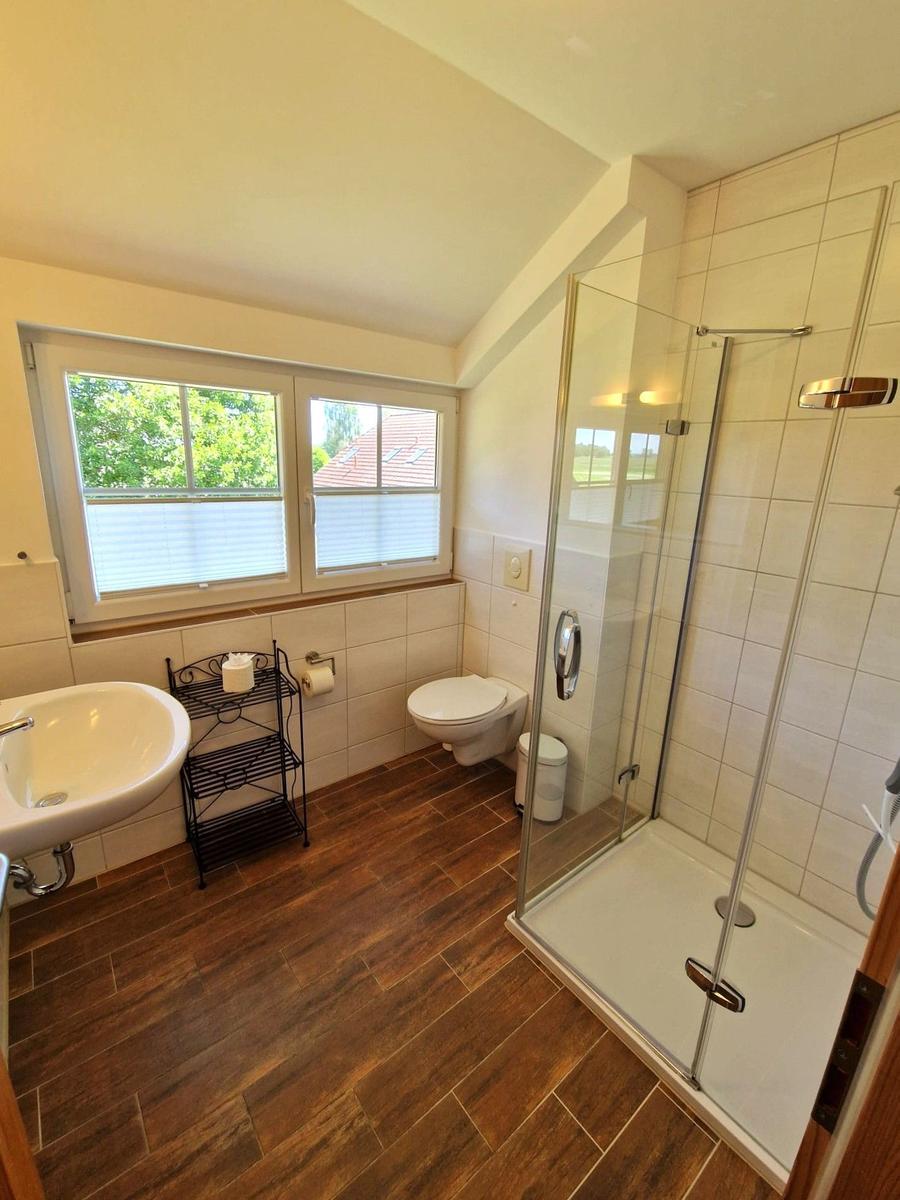 Badezimmer mit Dusche, Toilette und Waschbecken. Holzdielenboden und Fenster mit Blick.