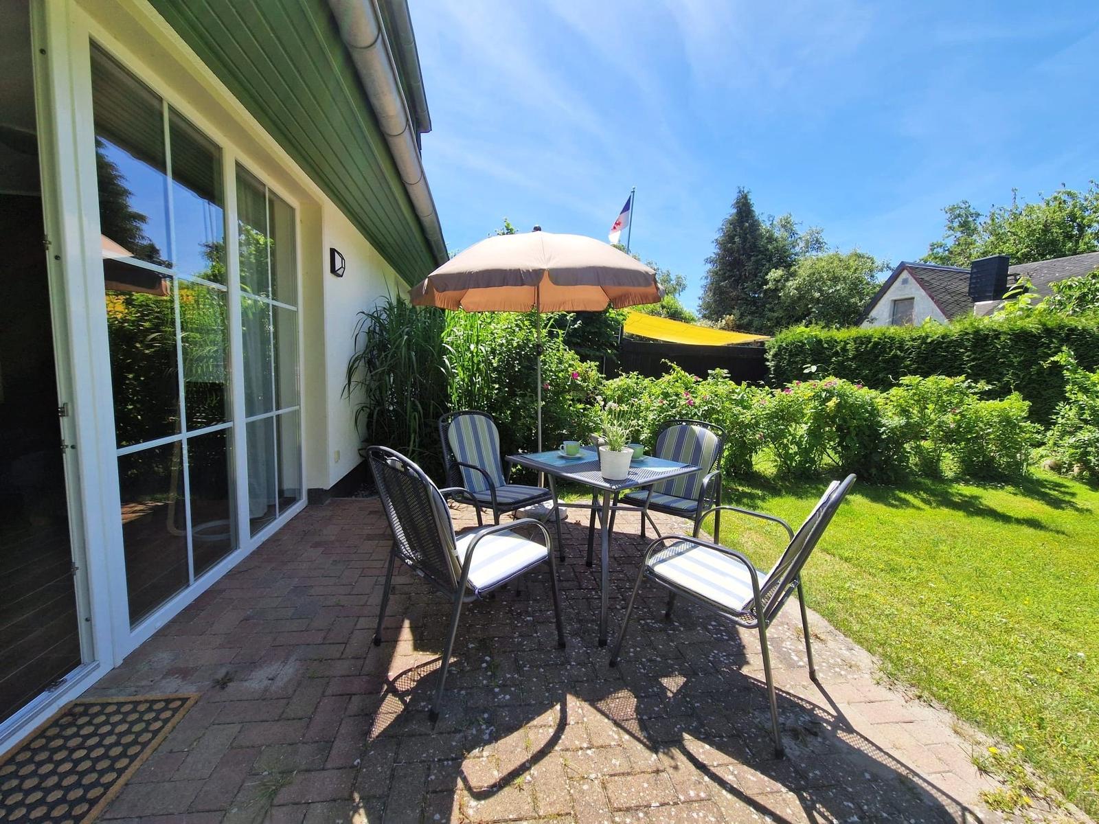 Terrasse mit Tisch, vier Stühlen und Sonnenschirm. Grünflächen und Hecke im Hintergrund.