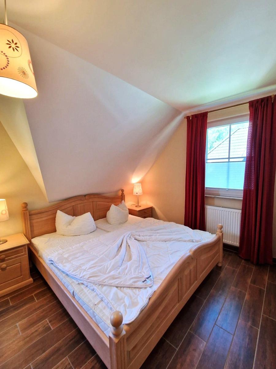 Schlafzimmer mit Holzbett, Nachttischen und Fenster mit roten Vorhängen.