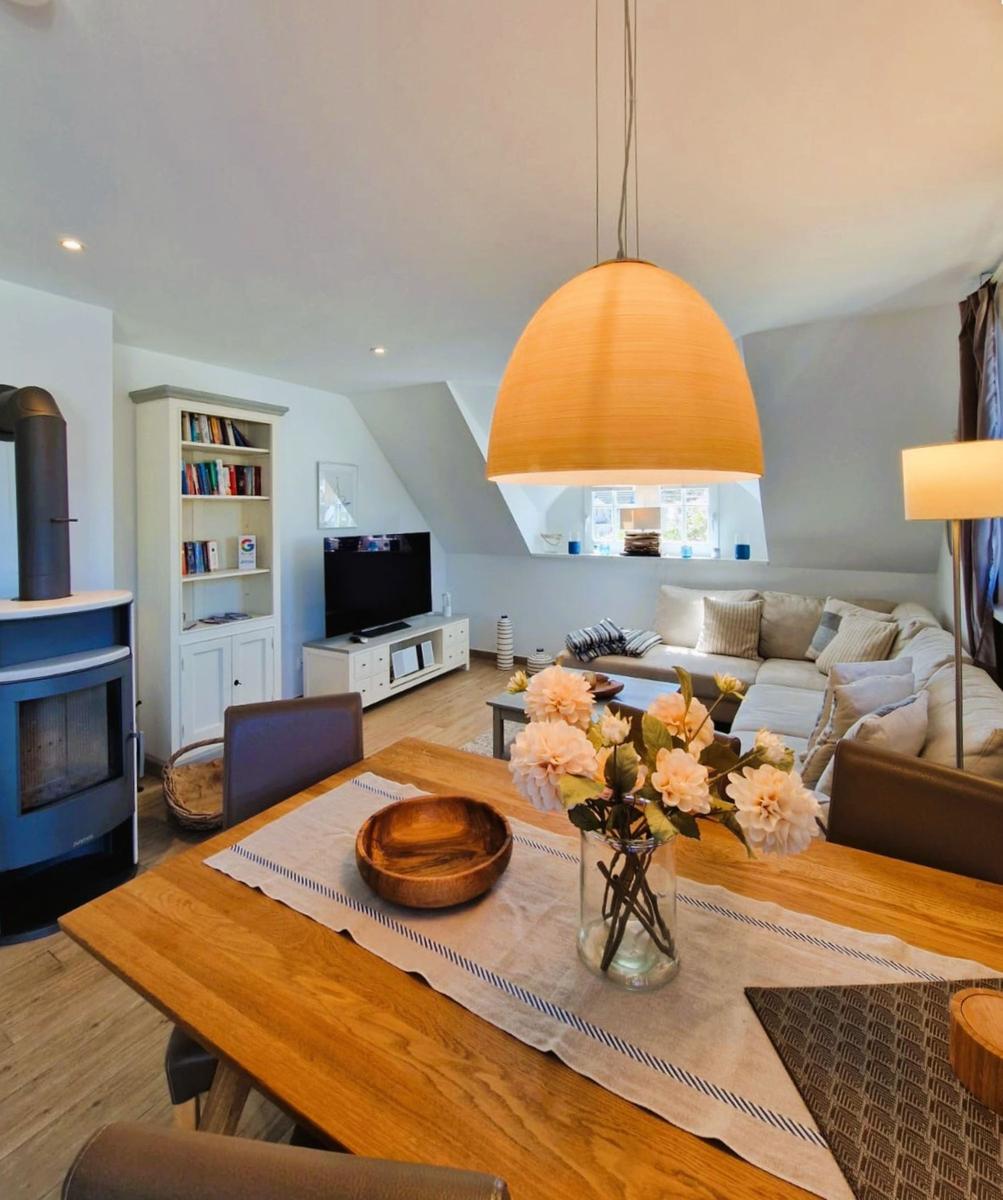 Essbereich mit Holztisch, Blumen und Deckenbeleuchtung. Im Hintergrund Wohnzimmer mit Sofa und Fernseher.