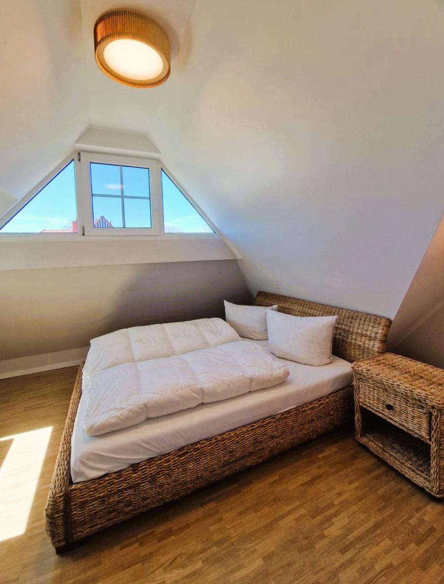 Schlafzimmer mit Dachschrägen, Bett und Nachttisch aus Rattan, Fenster mit Blick nach draußen.