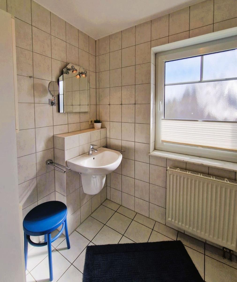 Badezimmer mit Waschbecken, Spiegel, Fenster und blauem Hocker.