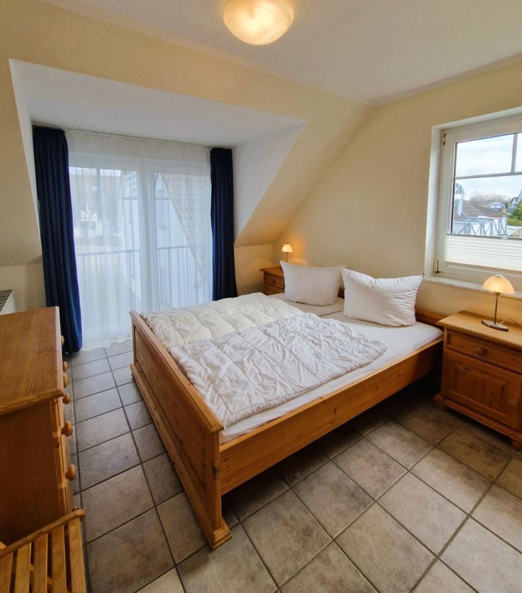 Schlafzimmer mit Holzbett, zwei Nachttischen und Fenstern. Bodenfliesen und Dachschräge.
