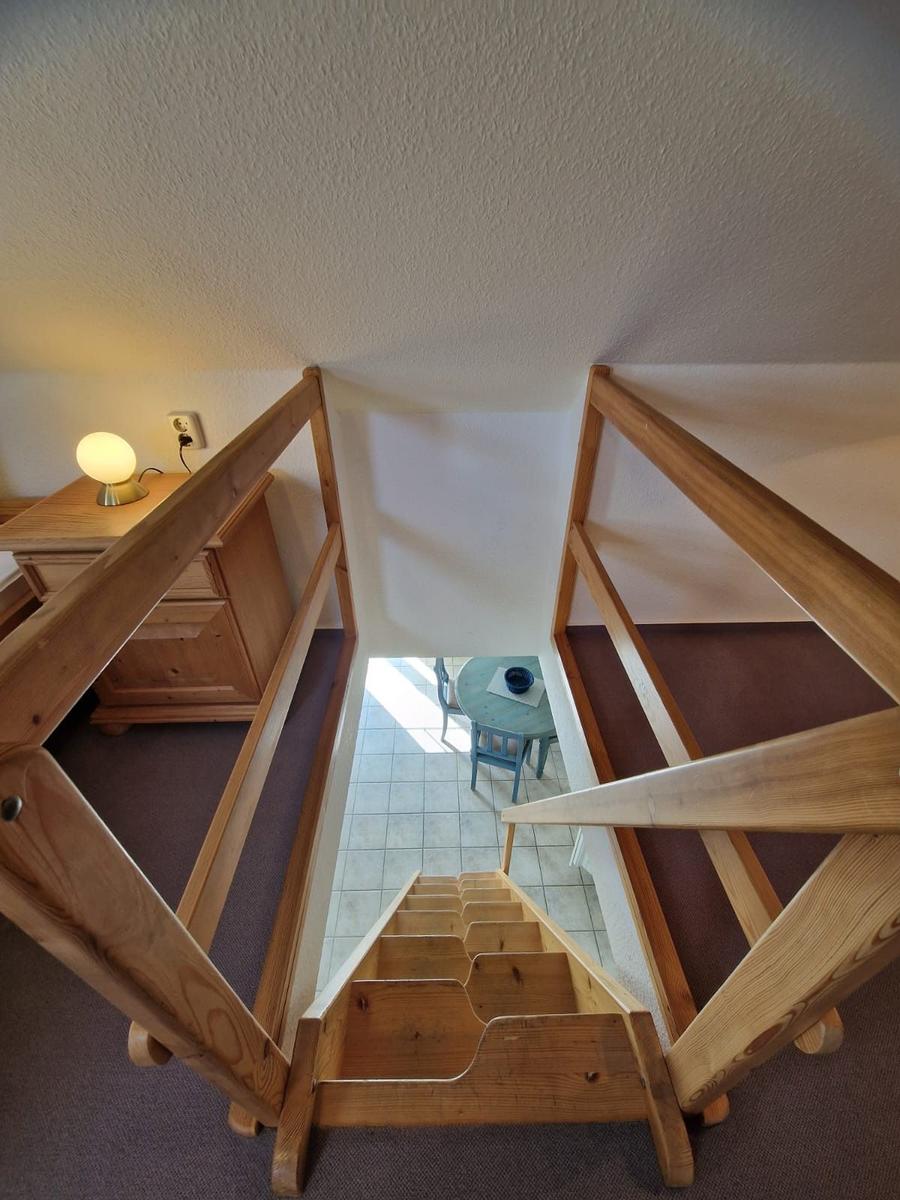 Holzstufen führen nach unten zu einem Tisch mit Stuhl. Links steht ein Nachttisch mit Lampe.