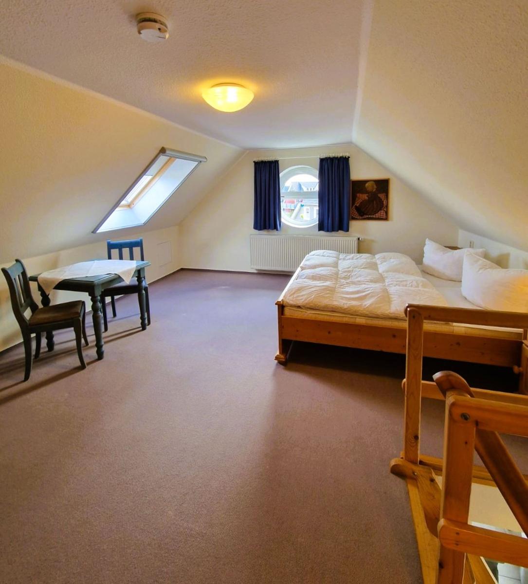 Zimmer mit Bett, Tisch, Stühlen, runden Fenster und Dachschrägen.