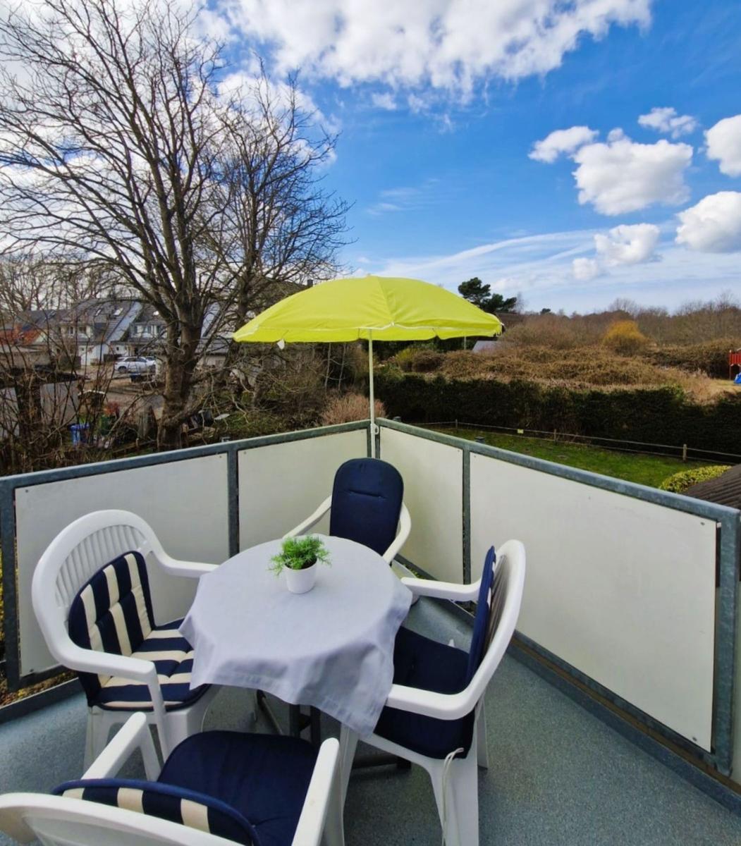Terrasse mit Tisch, vier Stühlen und gelbem Sonnenschirm. Blick auf Grünflächen und Bäume.