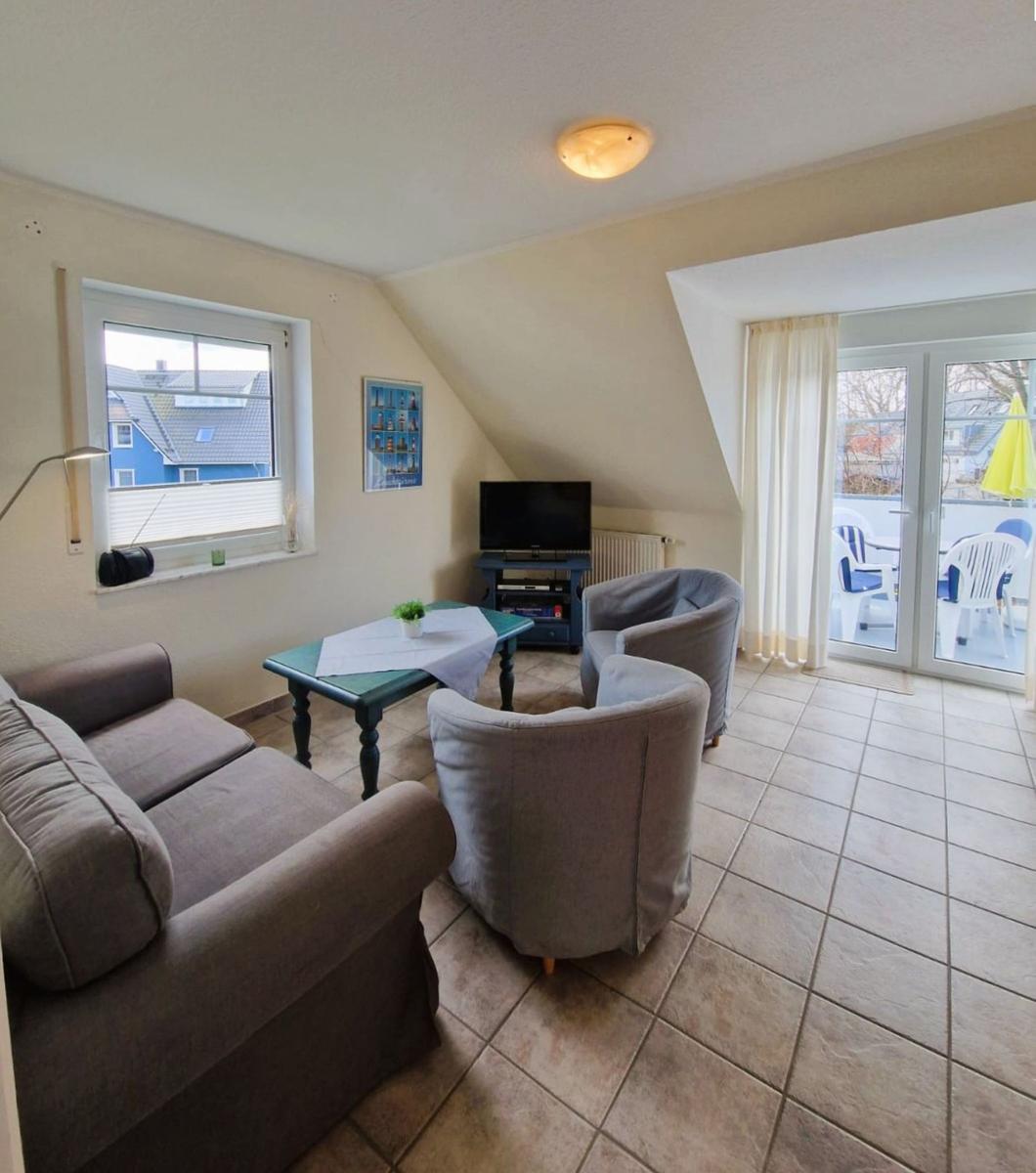 Wohnzimmer mit Sofa, zwei Sesseln, Tisch und TV. Durchgang zur Terrasse mit Gartenmöbeln.