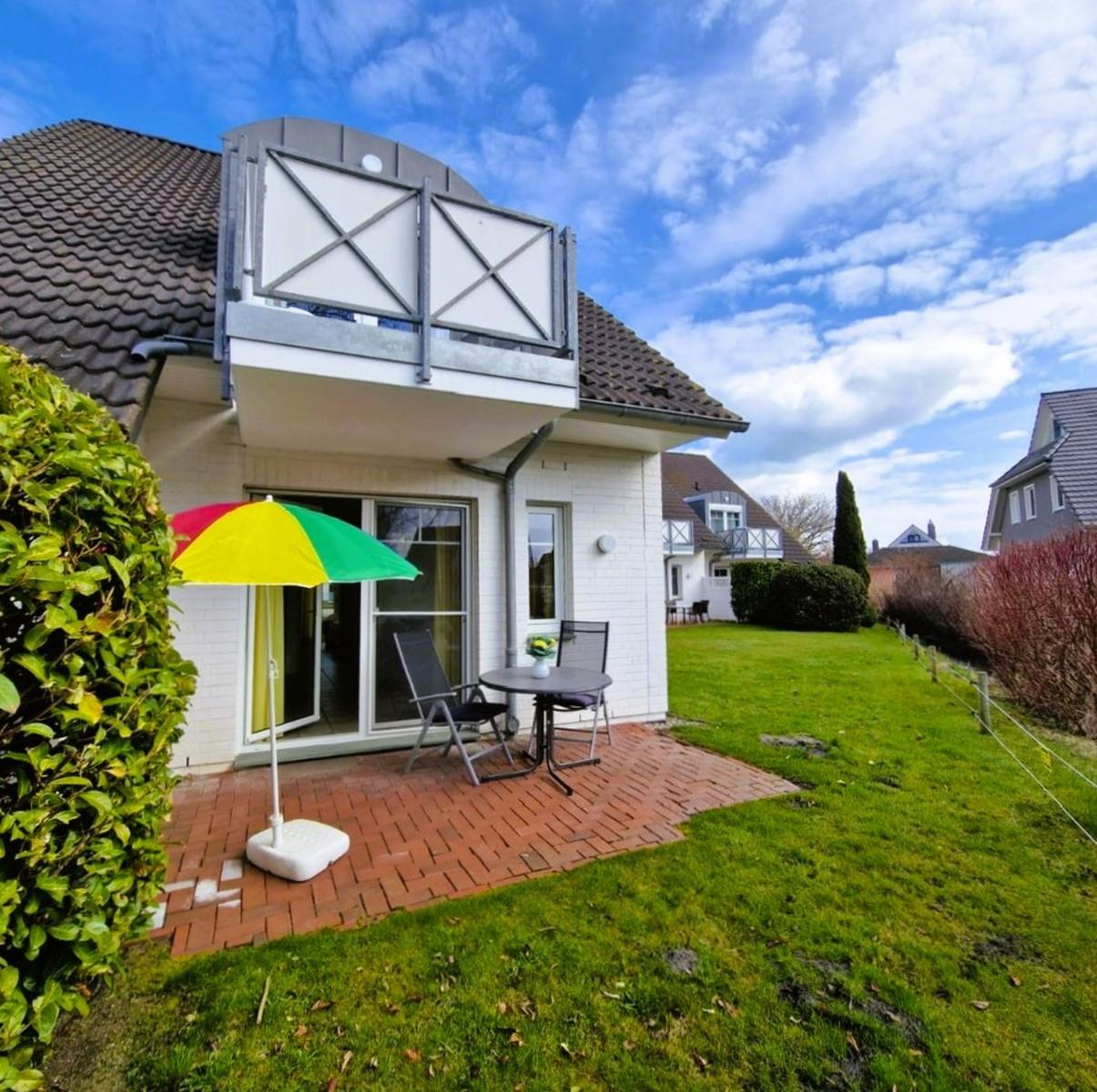 Haus mit Terrasse, Sitzgruppe und buntem Sonnenschirm. Grünflächen und Hinterhof sichtbar.