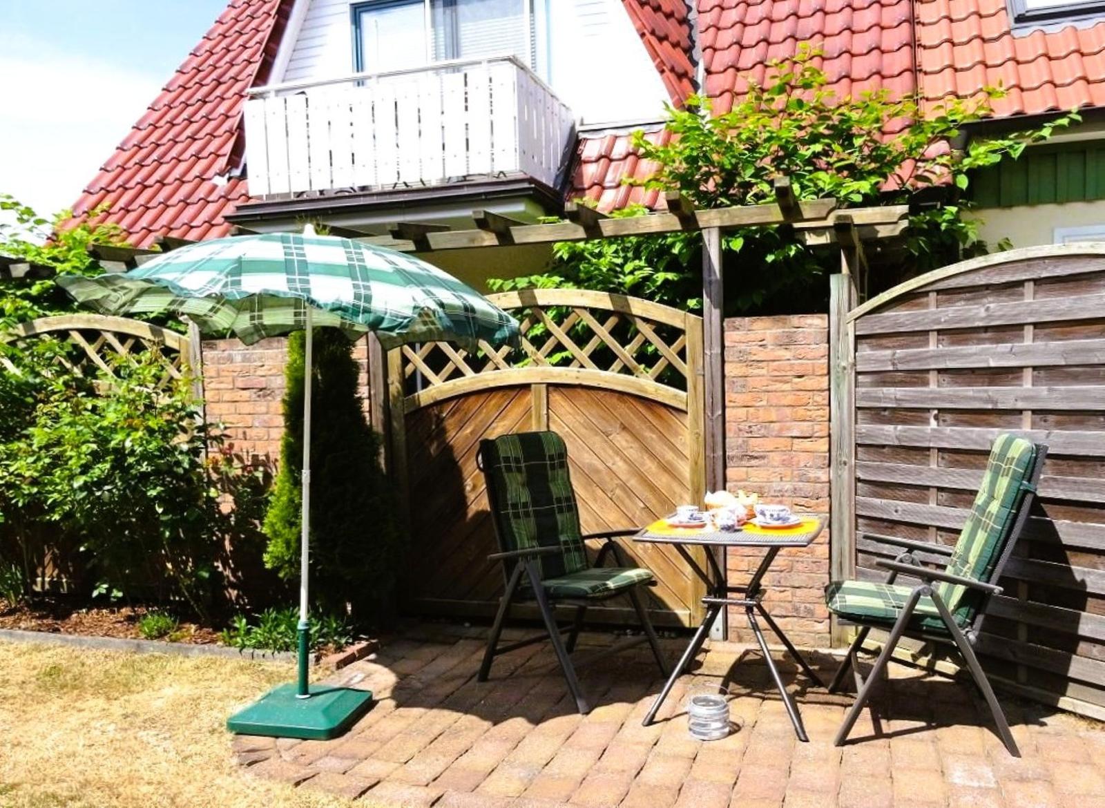 Terrace with table, two chairs, and umbrella. Background: wooden gate, fence, and house with red roof.