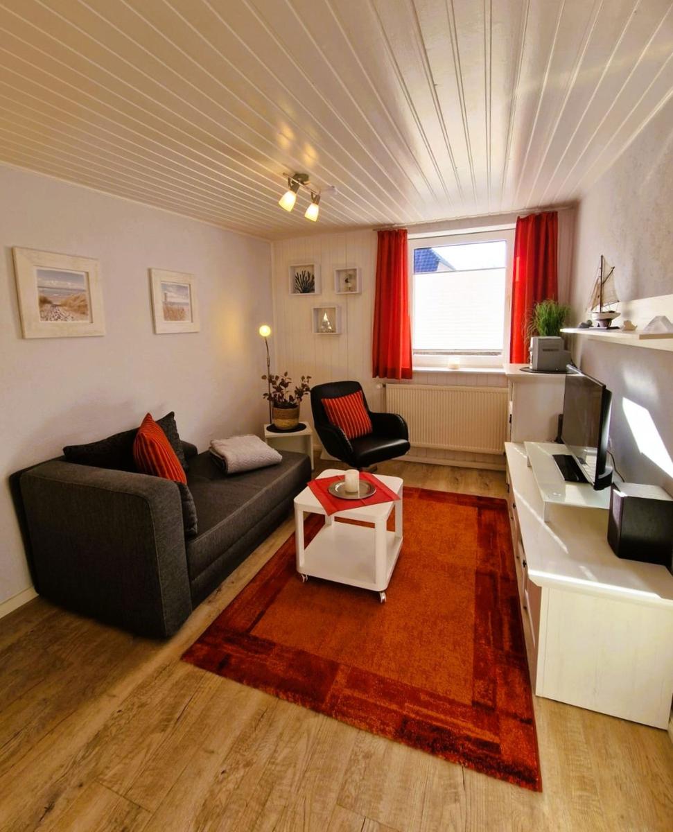 Living room with sofa, armchair, coffee table, and TV. Window with red curtains.
