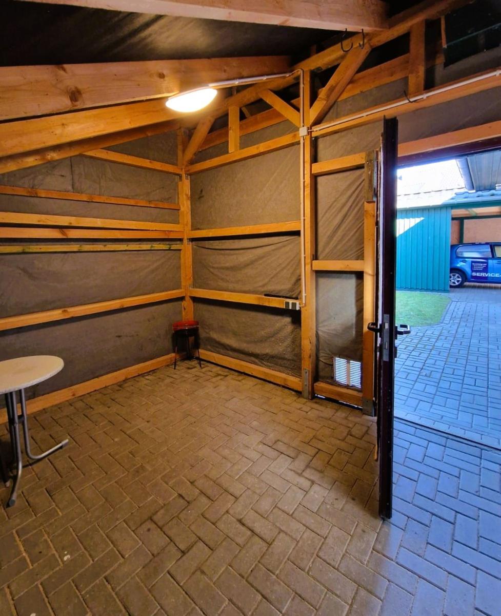 A room with wooden frame, brown floor, and an open door to the outside area.