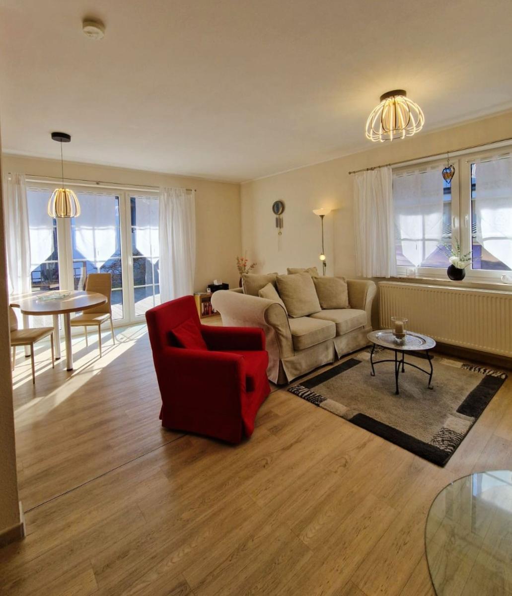 Living room with sofa, red armchair, and dining area. Wooden floor and large windows.