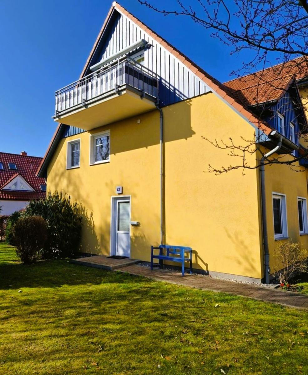Gelbes Haus mit Balkon, blauer Bank und grünem Garten vor der Tür.