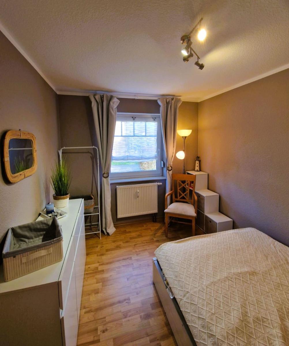 Schlafzimmer mit Bett, Fenster, Stuhl und Schrank. Holzfußboden und Wandspiegel.