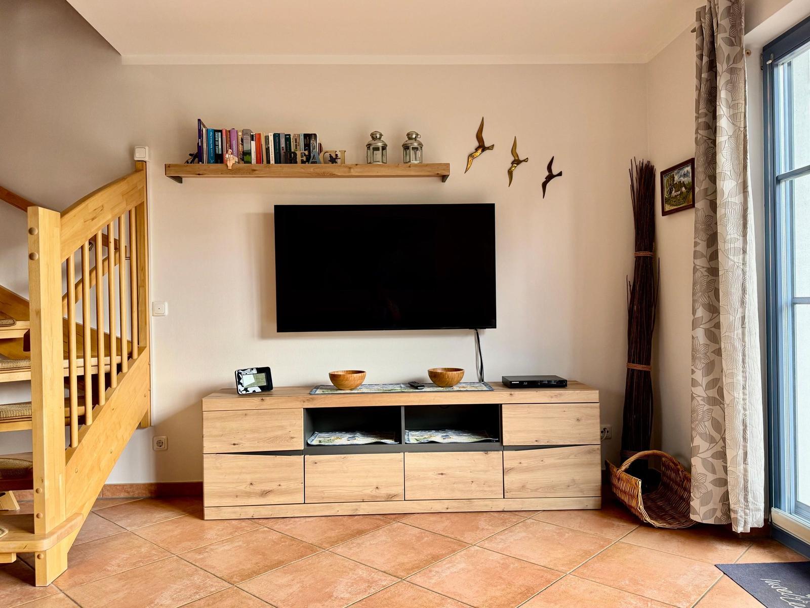 Wohnzimmer mit TV, Holzregal und Treppenaufgang.