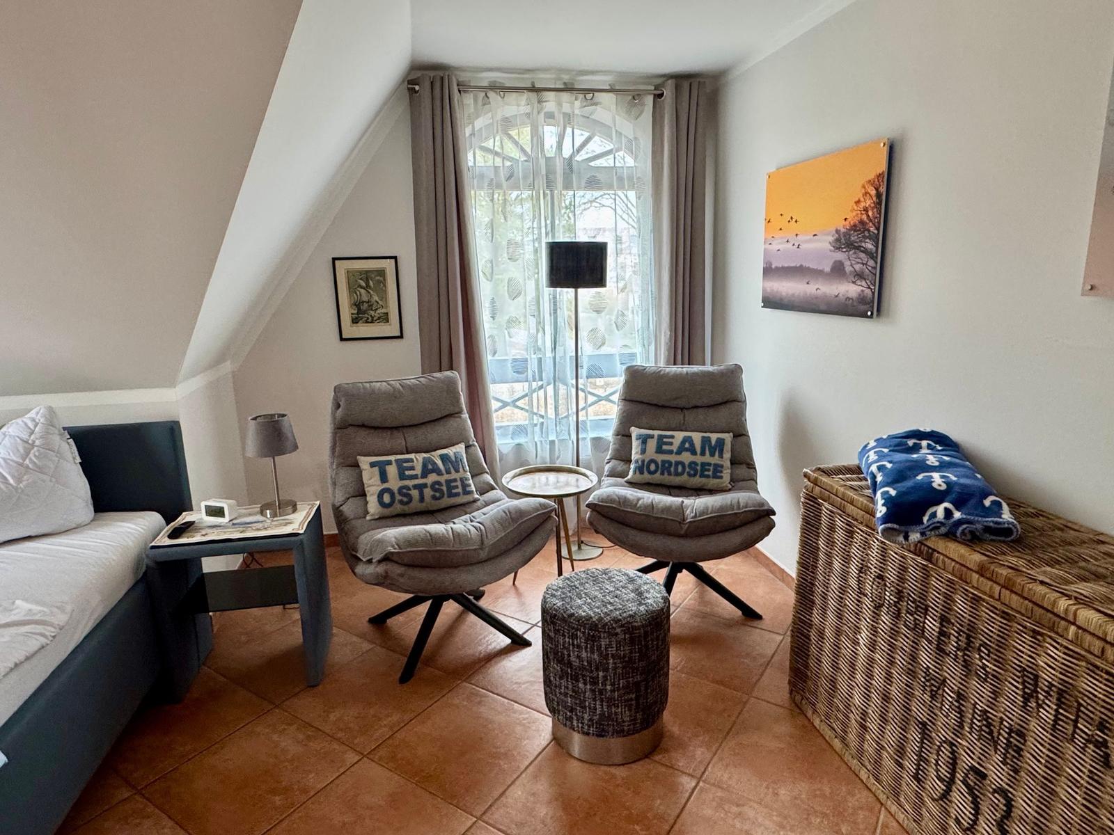 Zimmer mit zwei Sesseln, Bett und Korb. Fenster mit Vorhängen und Wandbild.