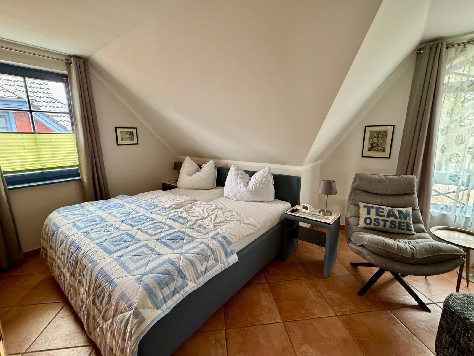 Schlafzimmer mit Doppelbett, Sessel und zwei Fenstern mit Vorhängen.