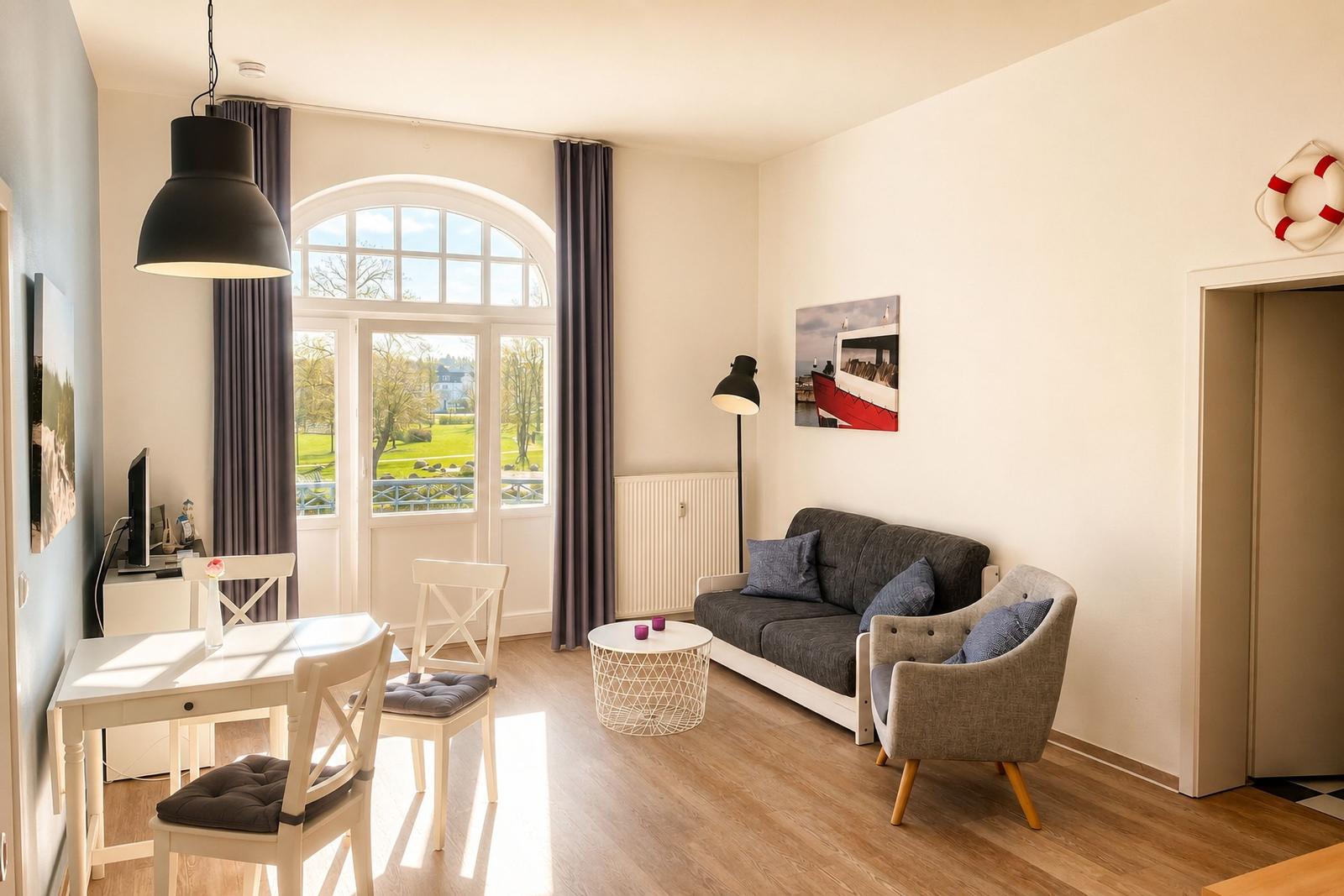 Wohn- und Essbereich mit Sofa, Stuhl und Tisch. Große Fensterfront mit Blick auf Grünfläche.