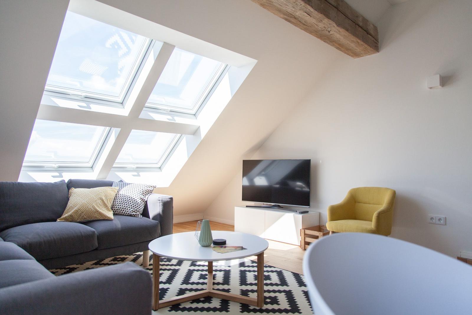 Wohnzimmer mit Sofa, Sessel und Dachfenstern. Fernseher und Couchtisch sichtbar.