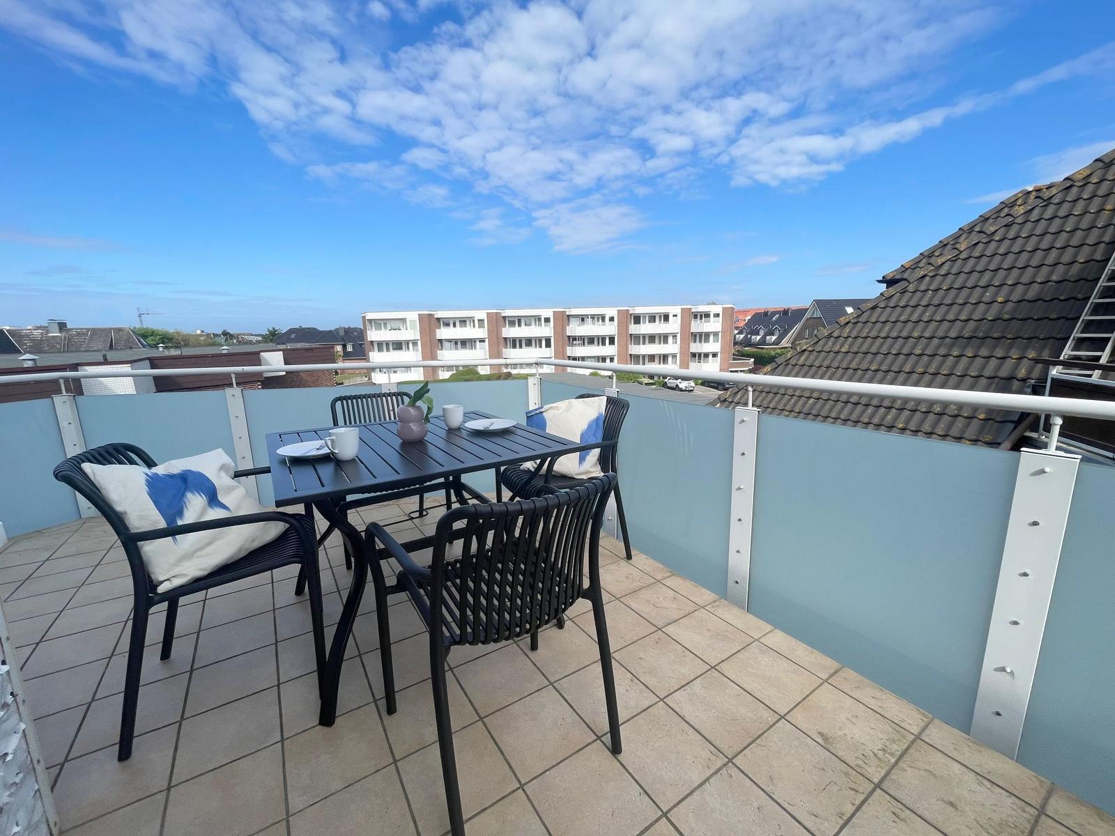 Terrasse mit Tisch, Stühlen und Blick auf Wohngebäude unter blauem Himmel.