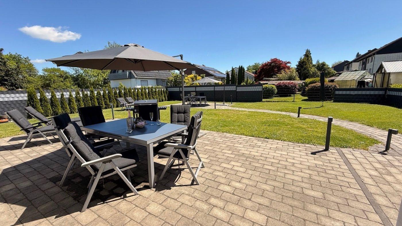 Terrasse mit Tisch, Stühlen und Sonnenschirm. Grasfläche und Zaun im Hintergrund.