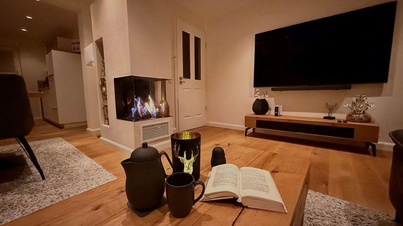 Wohnzimmer mit Kamin, Fernseher und Holzboden. Auf Tisch: Buch, Teekanne und Tassen.