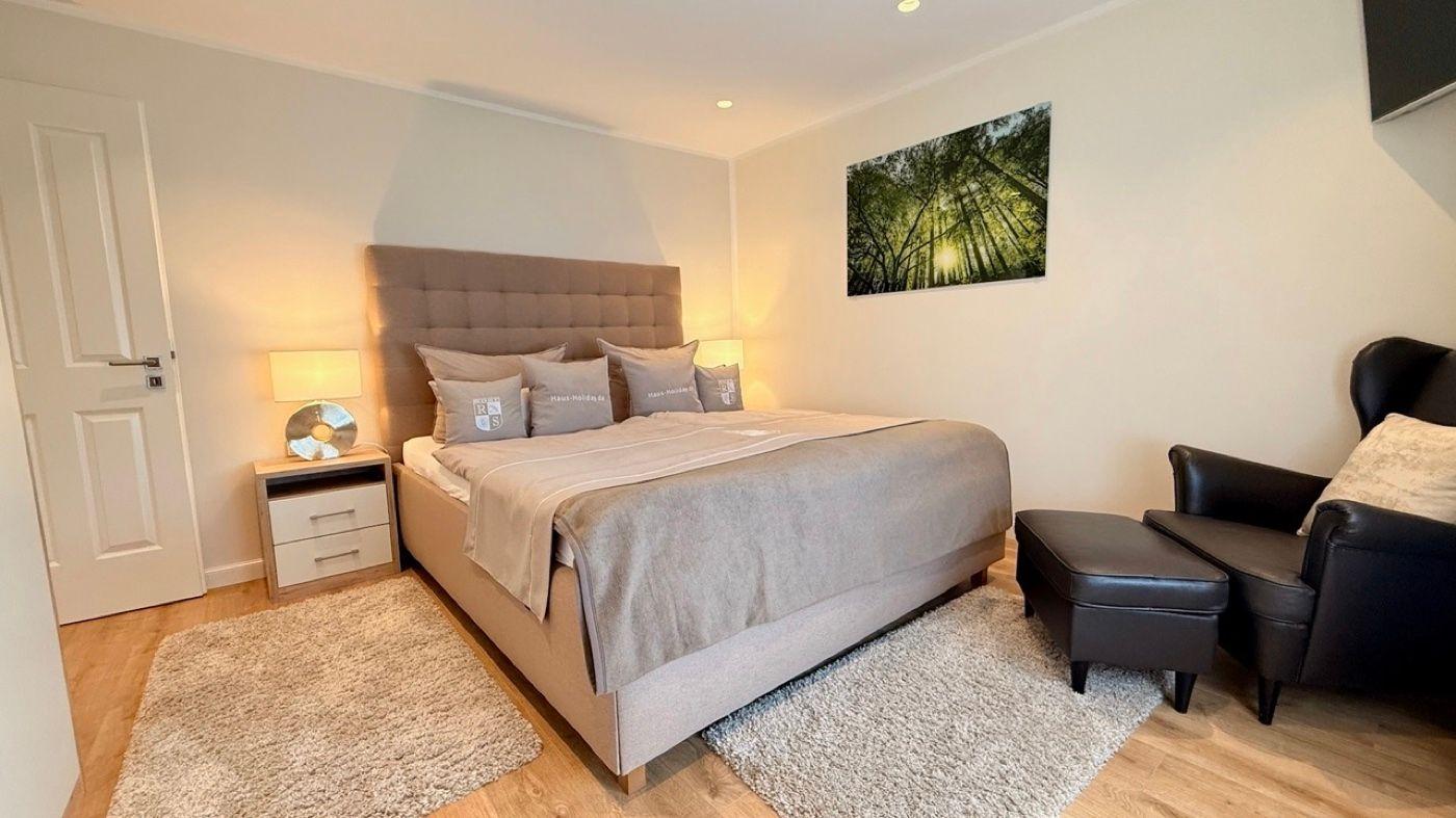 Schlafzimmer mit Bett, Nachttisch, Sessel und Wandbild.