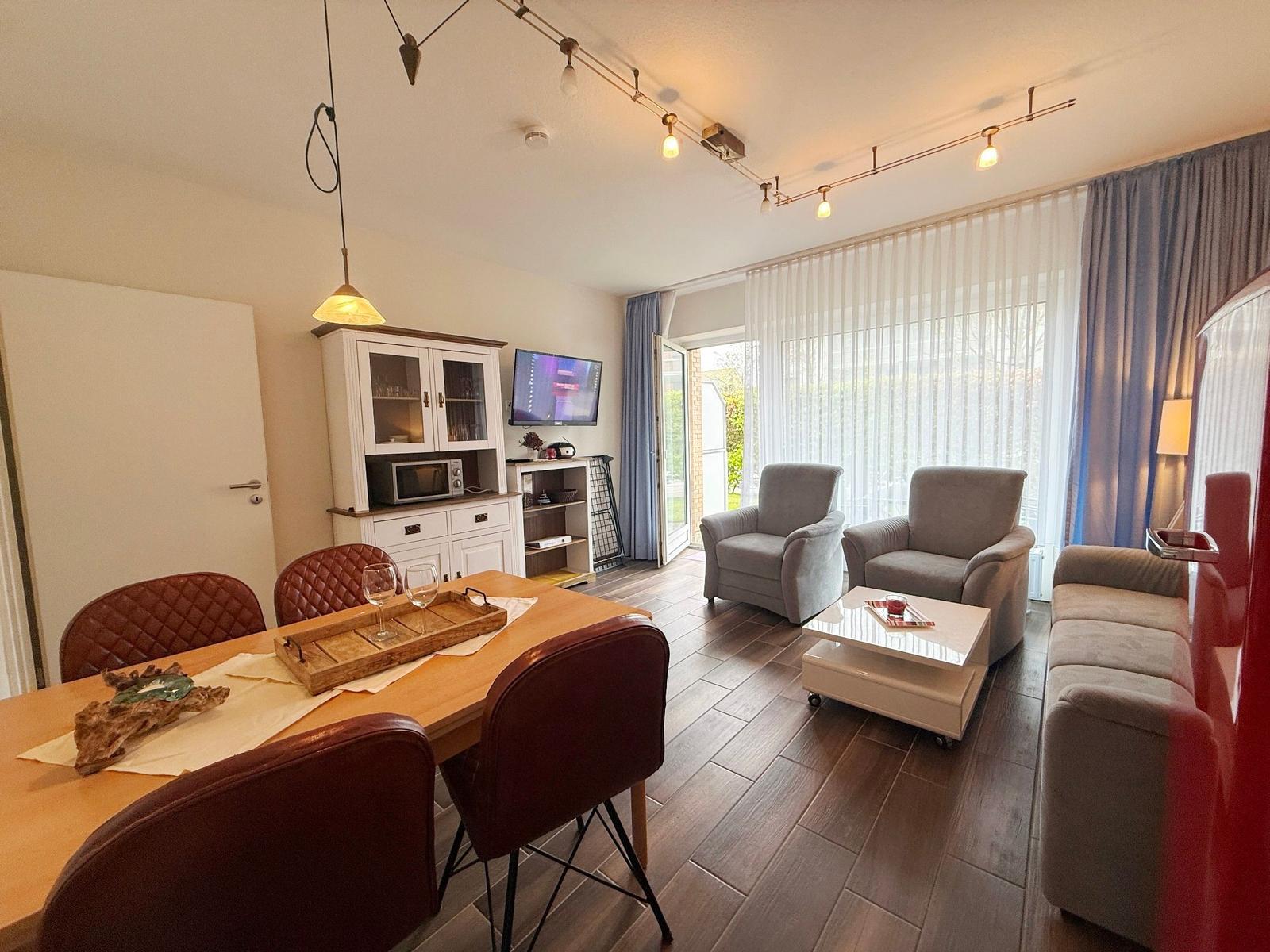 Esszimmer mit Tisch und Stühlen, Wohnbereich mit Sofa und Sesseln, TV und Fenster mit Vorhängen.