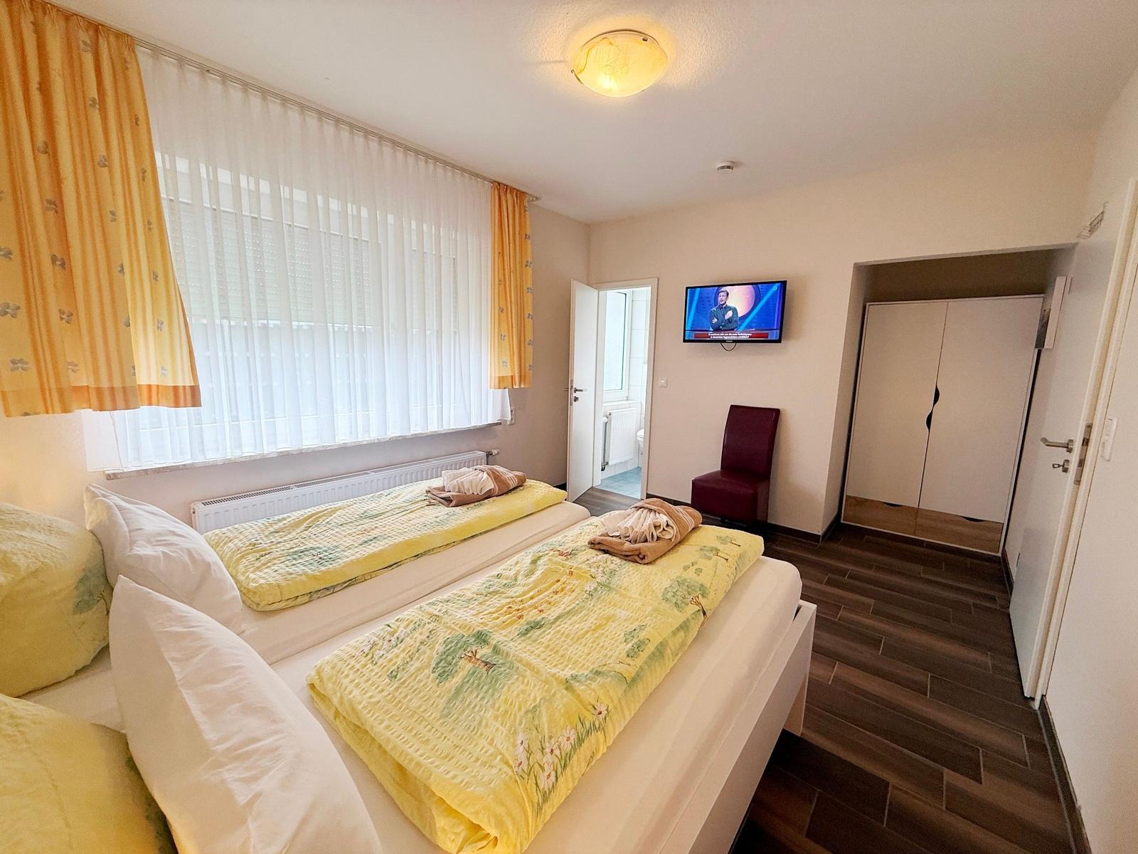 Zwei Betten mit gelben Bettwäschen, TV, Kleiderschrank und Fenster mit Vorhängen.