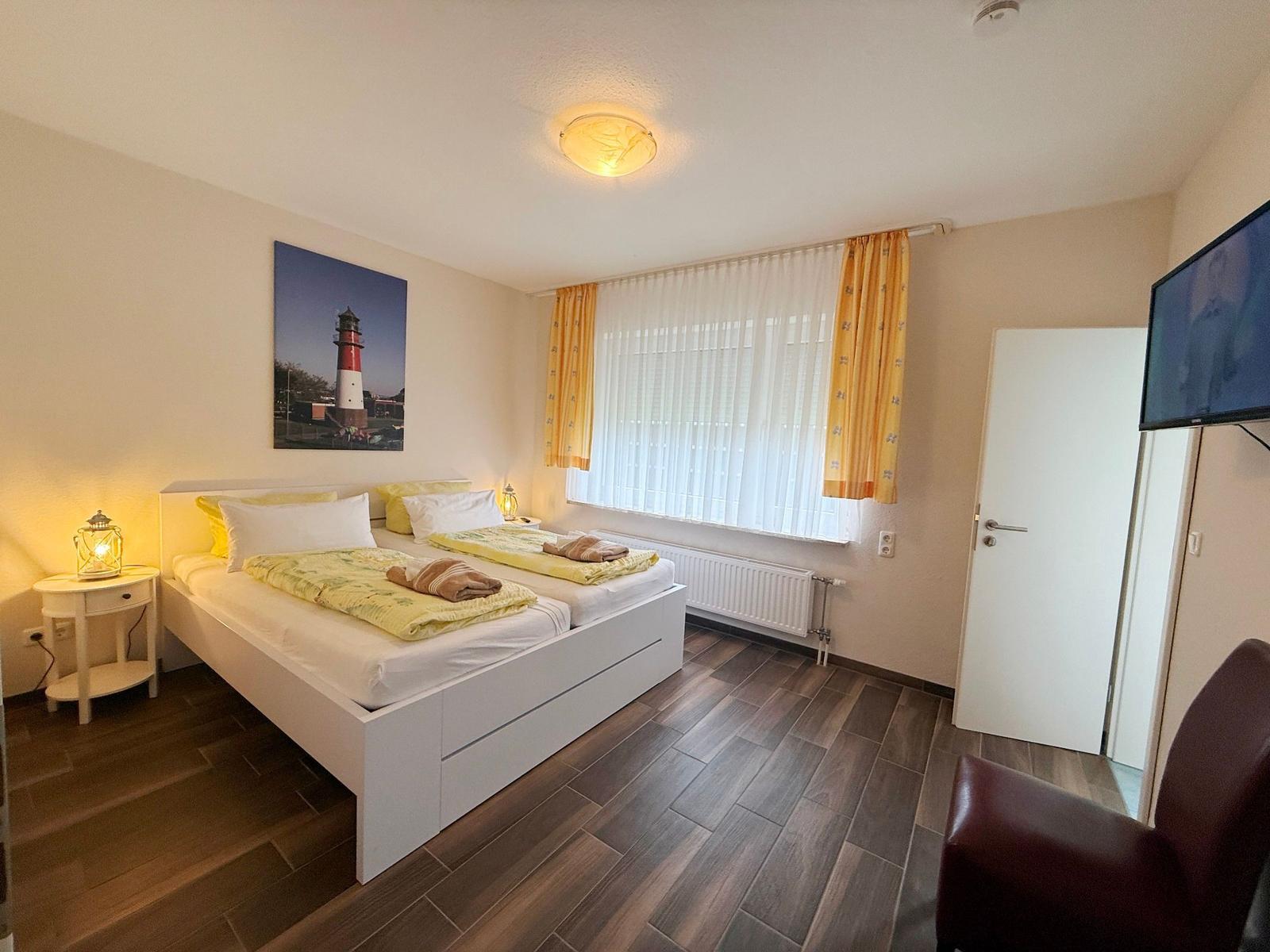 Schlafzimmer mit Doppelbett, Fenster, TV und Stuhl.