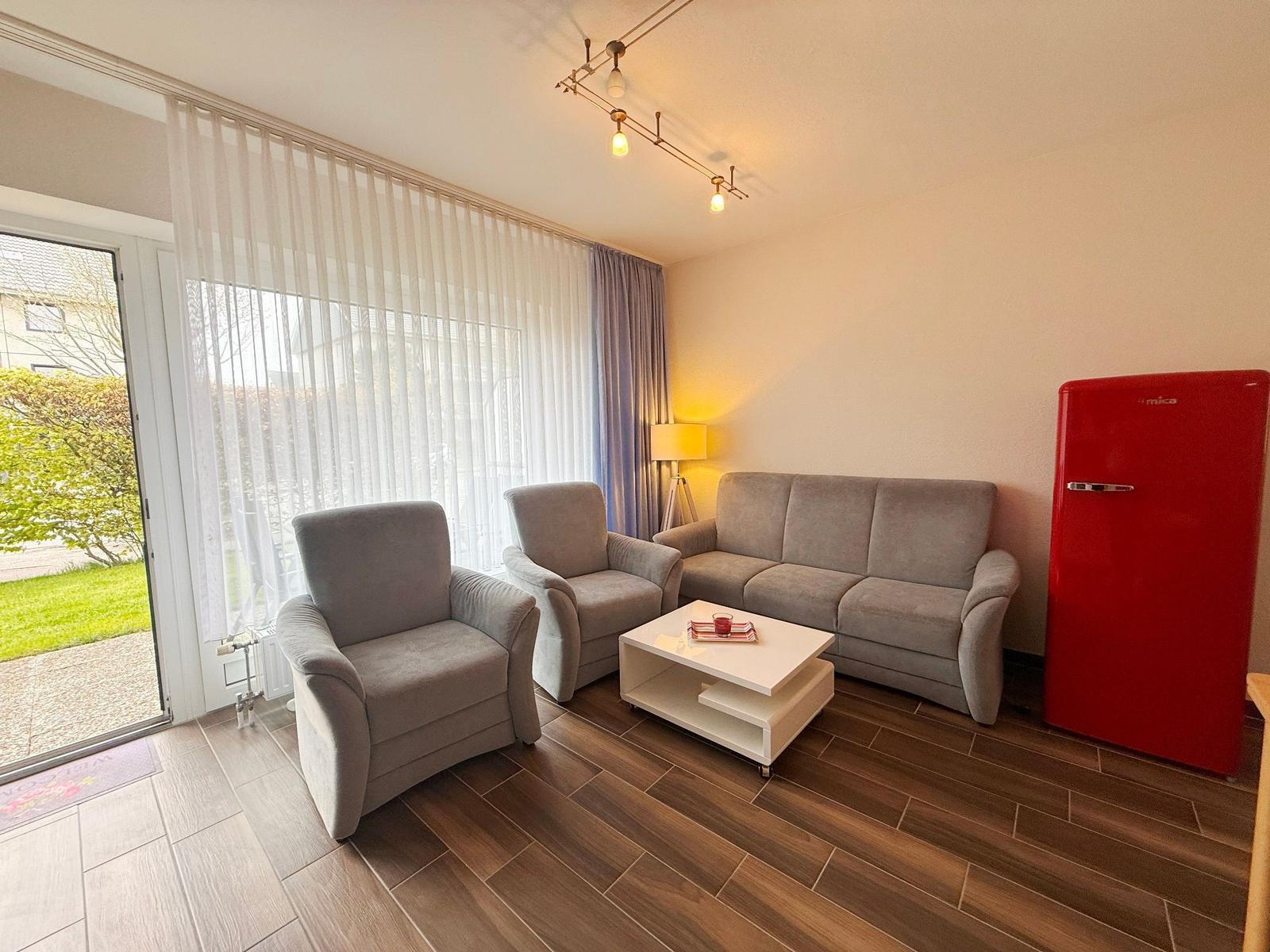 Wohnzimmer mit Sofa, zwei Sesseln, weißem Couchtisch und rotem Kühlschrank.